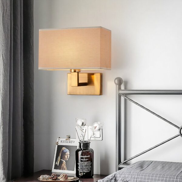 Wandlamp in bruine stof, met goudkleurige metalen beugel, naast een bed.