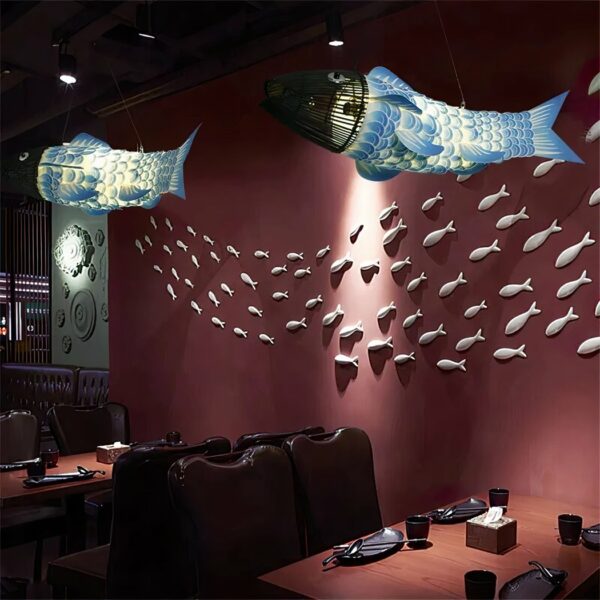 Restaurantzaal met twee kroonluchters in de vorm van grote vissen met blauwe en witte lichamen en koppen van zwarte houten stokken. Er zijn tafels gedekt en er is een rode muur op de achtergrond met veel vissen die aan de muur hangen als een school vissen in de zee.