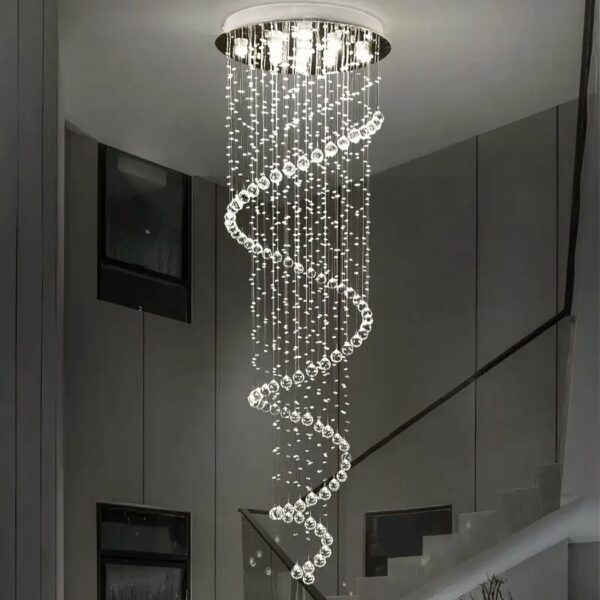 Plafondkroonluchter in de vorm van een spiraal van traanvormige ophangingen. Wit licht dat verlicht wordt door verschillende LED's in de ronde zilveren beugel die aan het plafond bevestigd is. Daarachter een trap met zwarte leuning en schilderijen aan de muur.